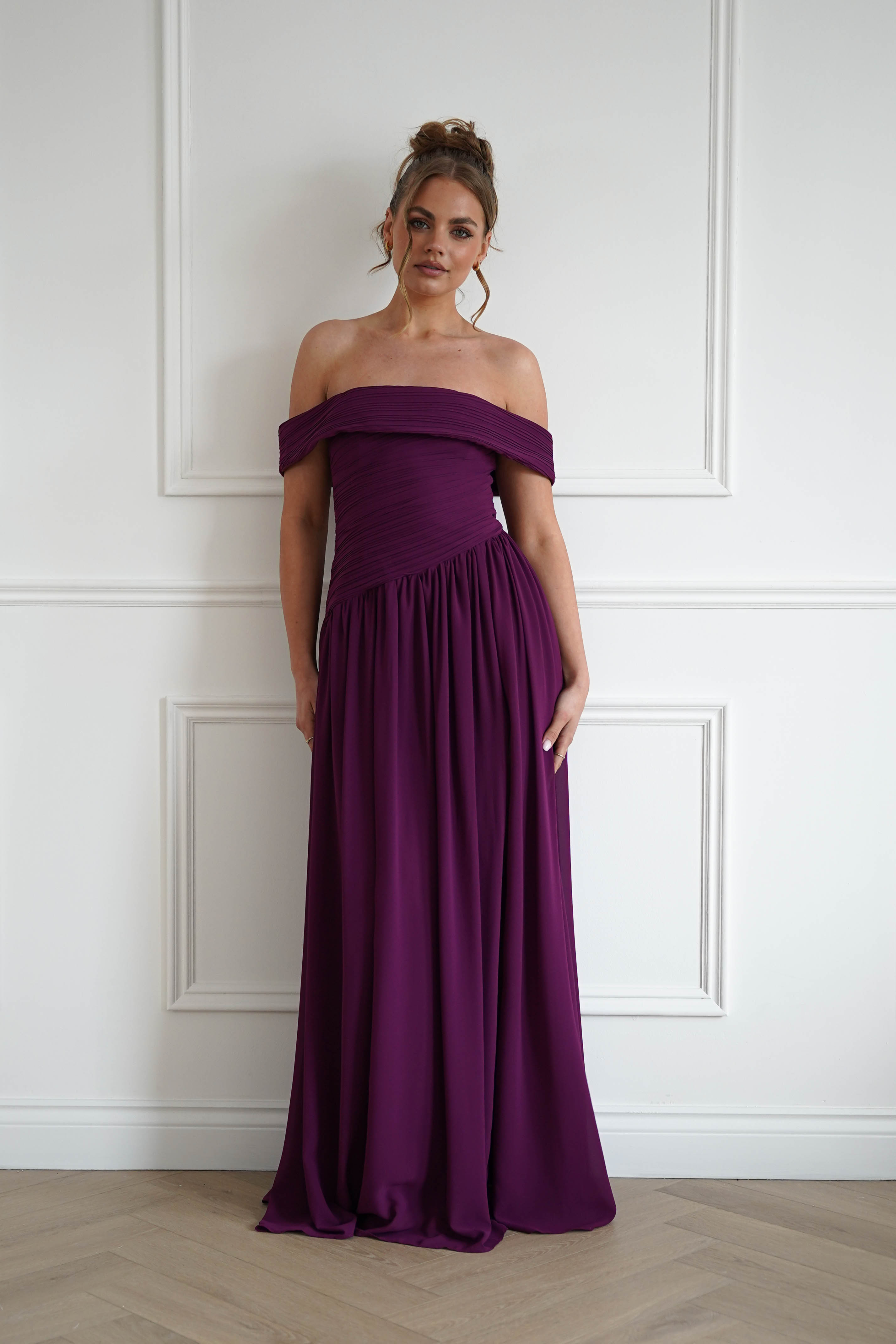 Woman wearing a plum off-the-shoulder chiffon maxi dress with a ruched bodice, gathered waist, and flowing floor-length skirt, styled for weddings, bridesmaid events, and formal occasions.