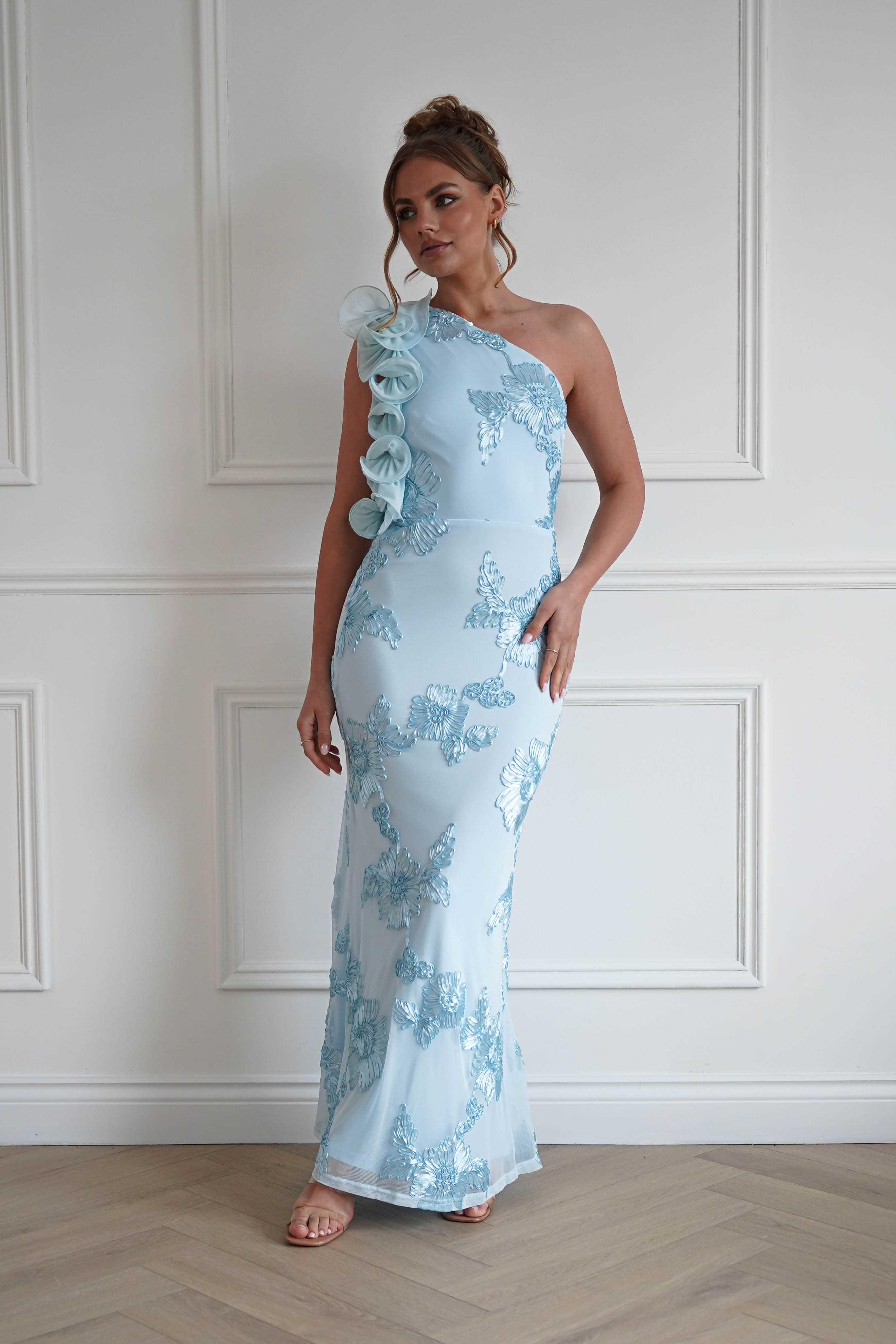 Woman wearing a light blue one-shoulder bodycon maxi dress with floral embroidered detailing and statement ruffle shoulder accents, ideal for weddings, races, and formal occasions.