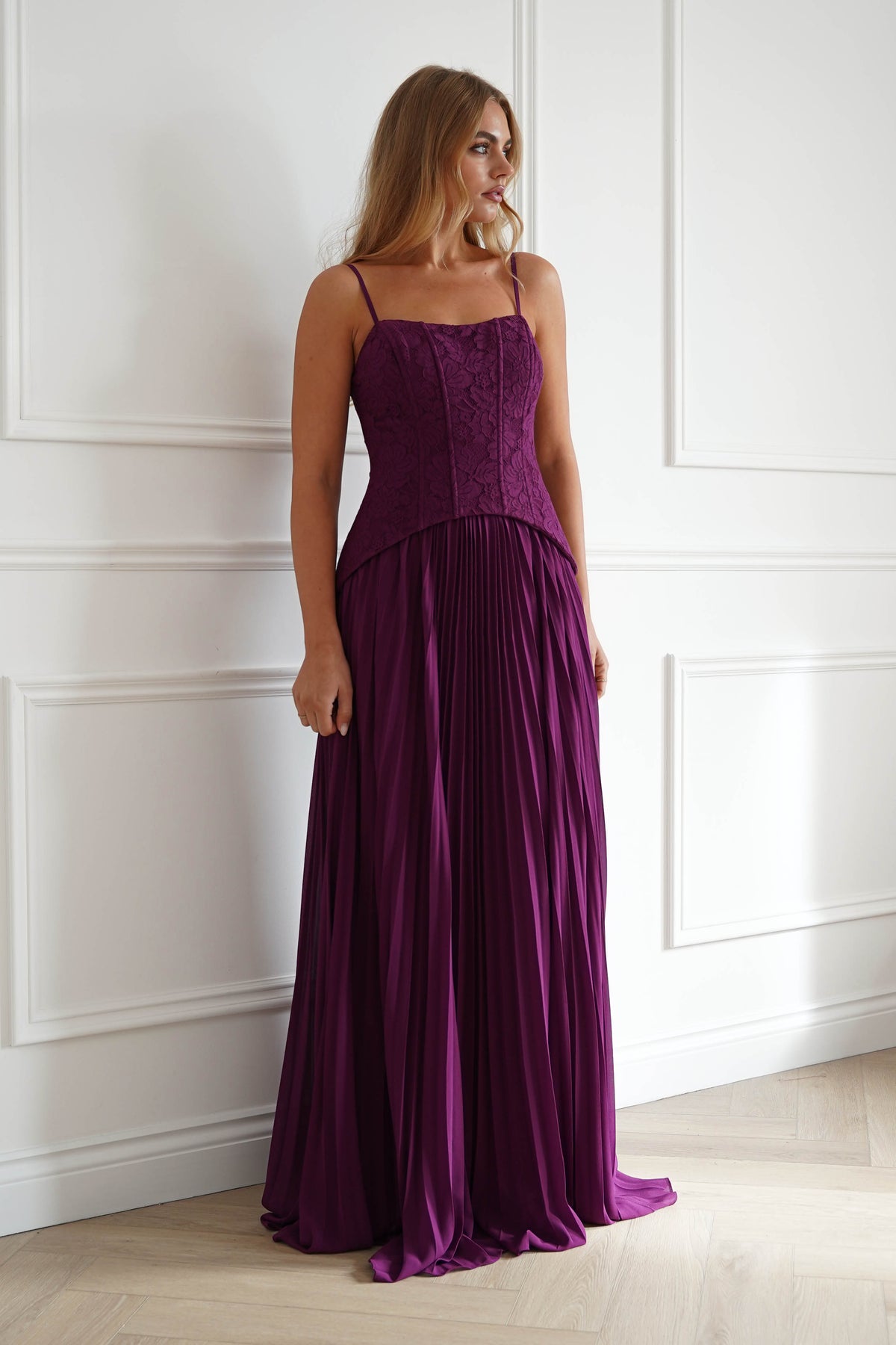A woman modeling a floor-length formal gown in a rich deep purple color, standing against a white paneled wall.