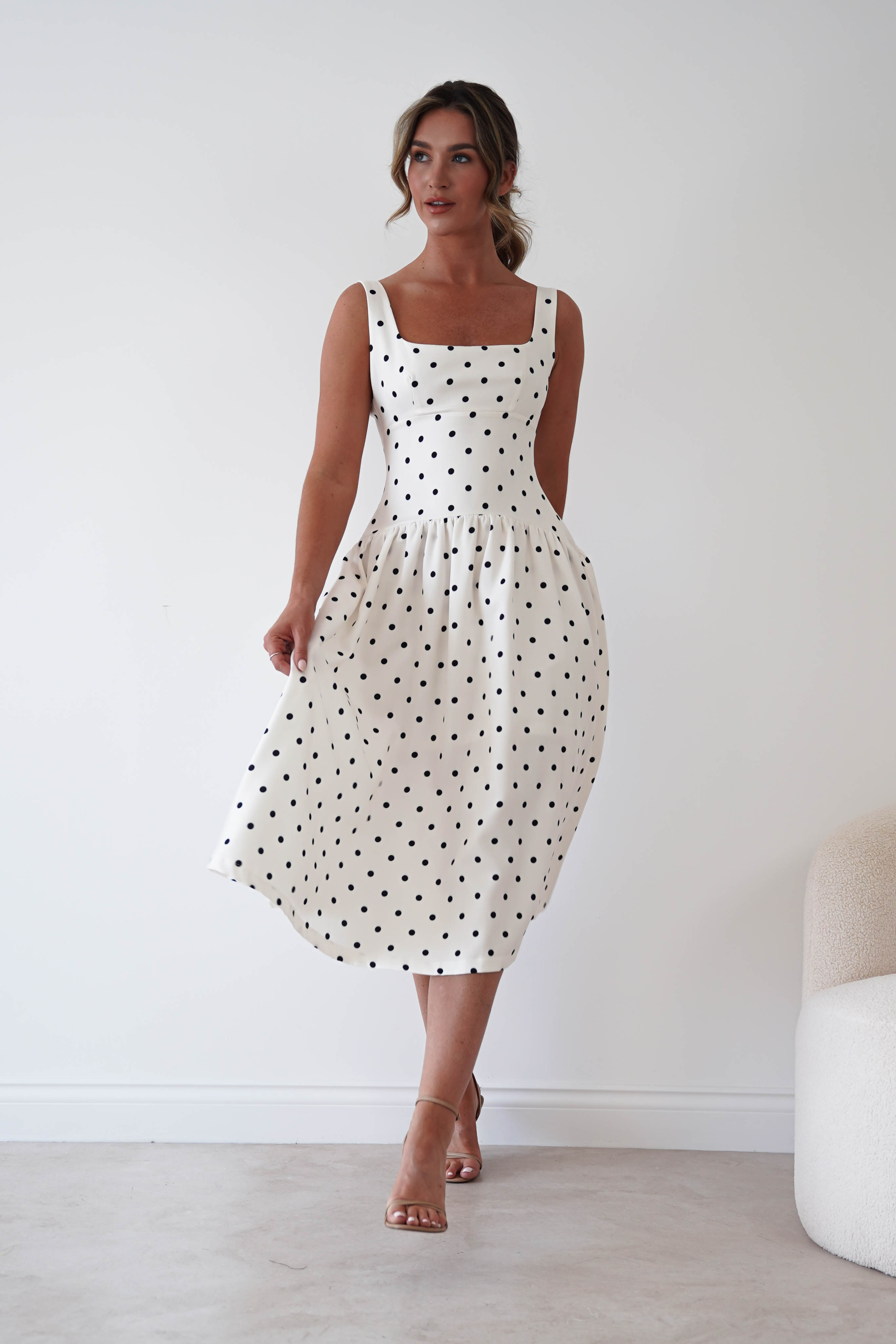 A woman modeling a sleeveless midi dress with a black polka dot pattern on a white background, standing in a minimalist room with a light-colored floor and wall.