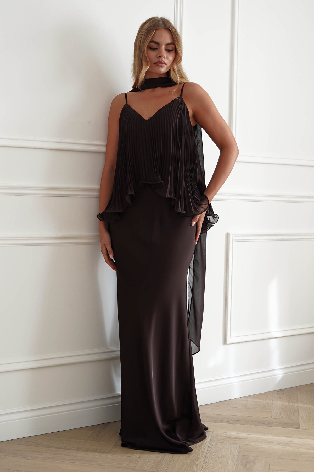 A woman models a dark brown, floor-length evening gown with a pleated halter top and a sheer draped overlay, standing against a white paneled wall.