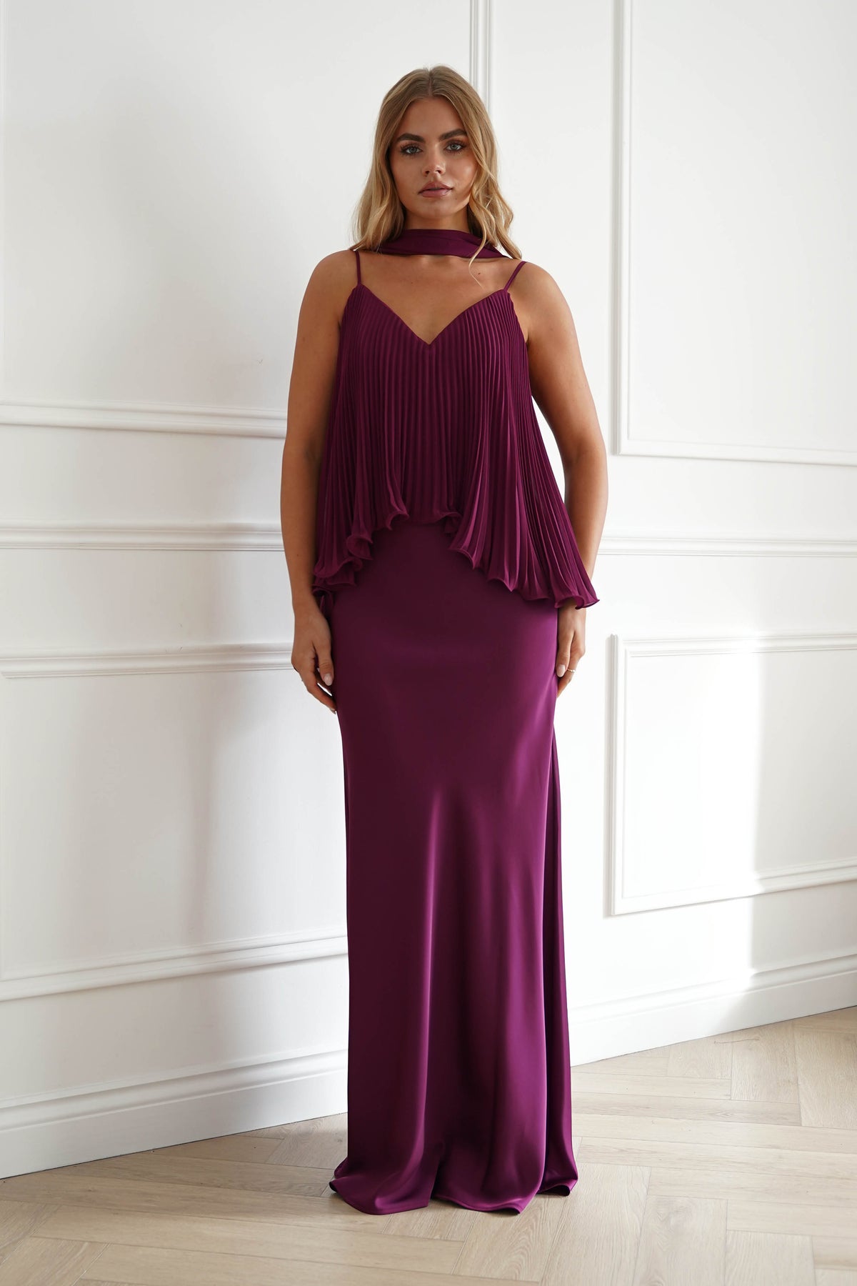 A woman modeling a floor-length plum purple gown with a pleated halter-style top and a flowing skirt, standing in a bright room with white paneled walls and light wood flooring.