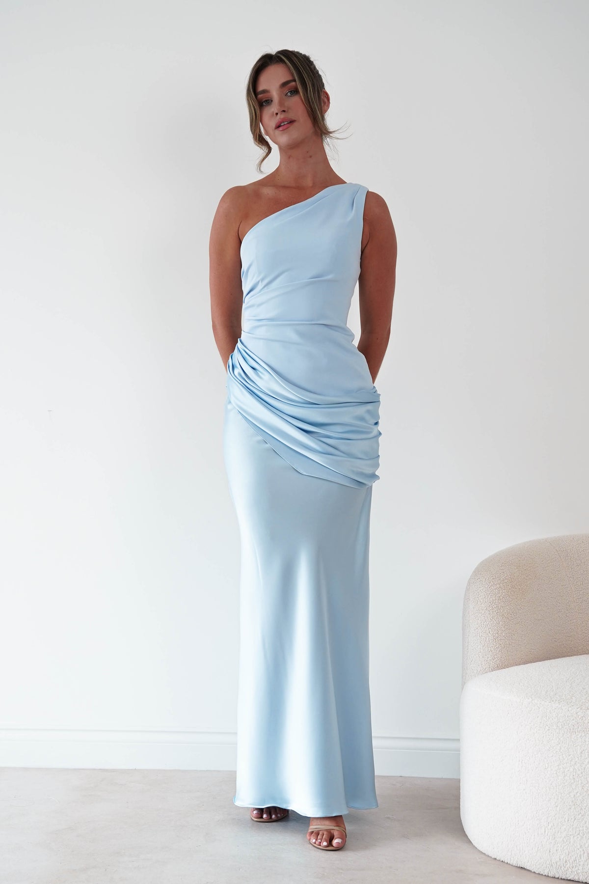 A woman modeling a light blue one-shoulder maxi dress with ruched detailing at the waist, standing in a minimalist room with a white wall and a cream-colored chair partially visible on the right.