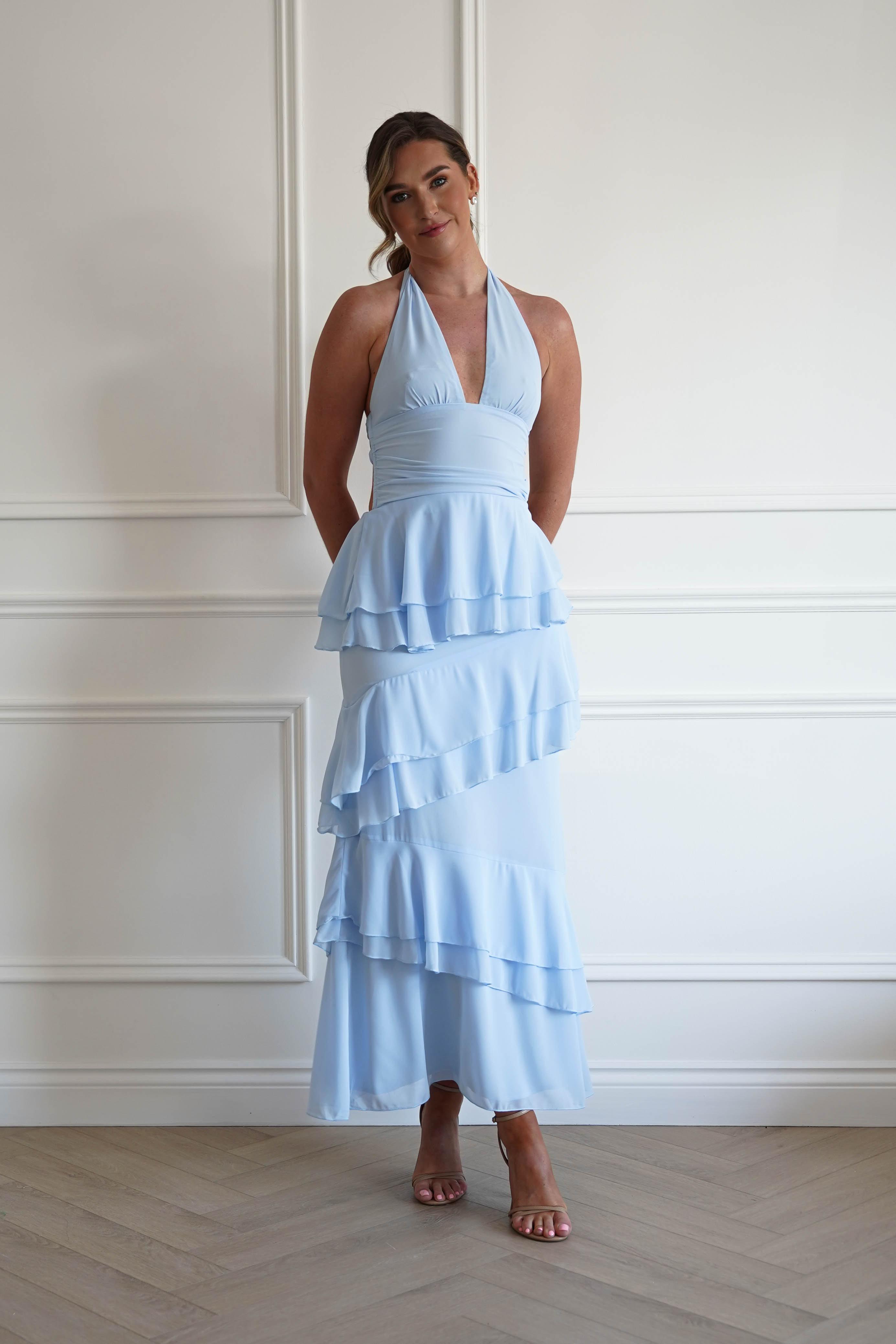 Woman wearing a light blue ruffled dress against a white paneled wall.
