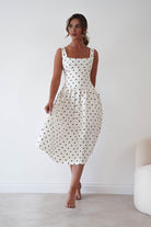 A woman modeling a sleeveless midi dress with a black polka dot pattern on a white background, standing in a minimalist room with a light-colored floor and wall.